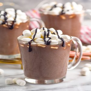 Glass mug of creamy crock pot hot chocolate topped with mini marshmallows and drizzled chocolate syrup, perfect Christmas drink.